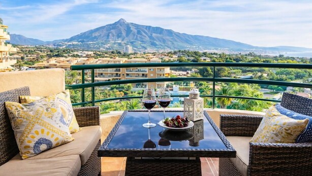 Un balcon agrémenté de meubles en osier, une table avec des verres à vin et une assiette de fruits, et une vue sur la montagne.