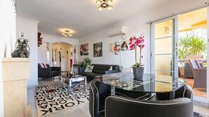 Un salon moderne avec une table à manger au plateau en verre, des fauteuils en cuir noir, un canapé noir et une table basse. Une grande fenêtre offre une vue sur la verdure à l’extérieur.