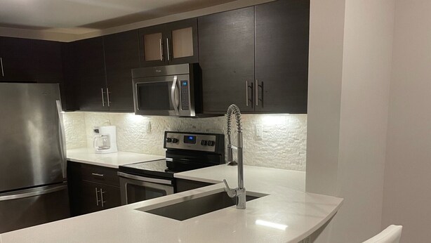 A modern kitchen with dark cabinets, stainless steel appliances, a white countertop, and a bar area with two white bar stools. - Luxury Prime Brickell Condo, Pool, Gym, Free Parking (Miami)