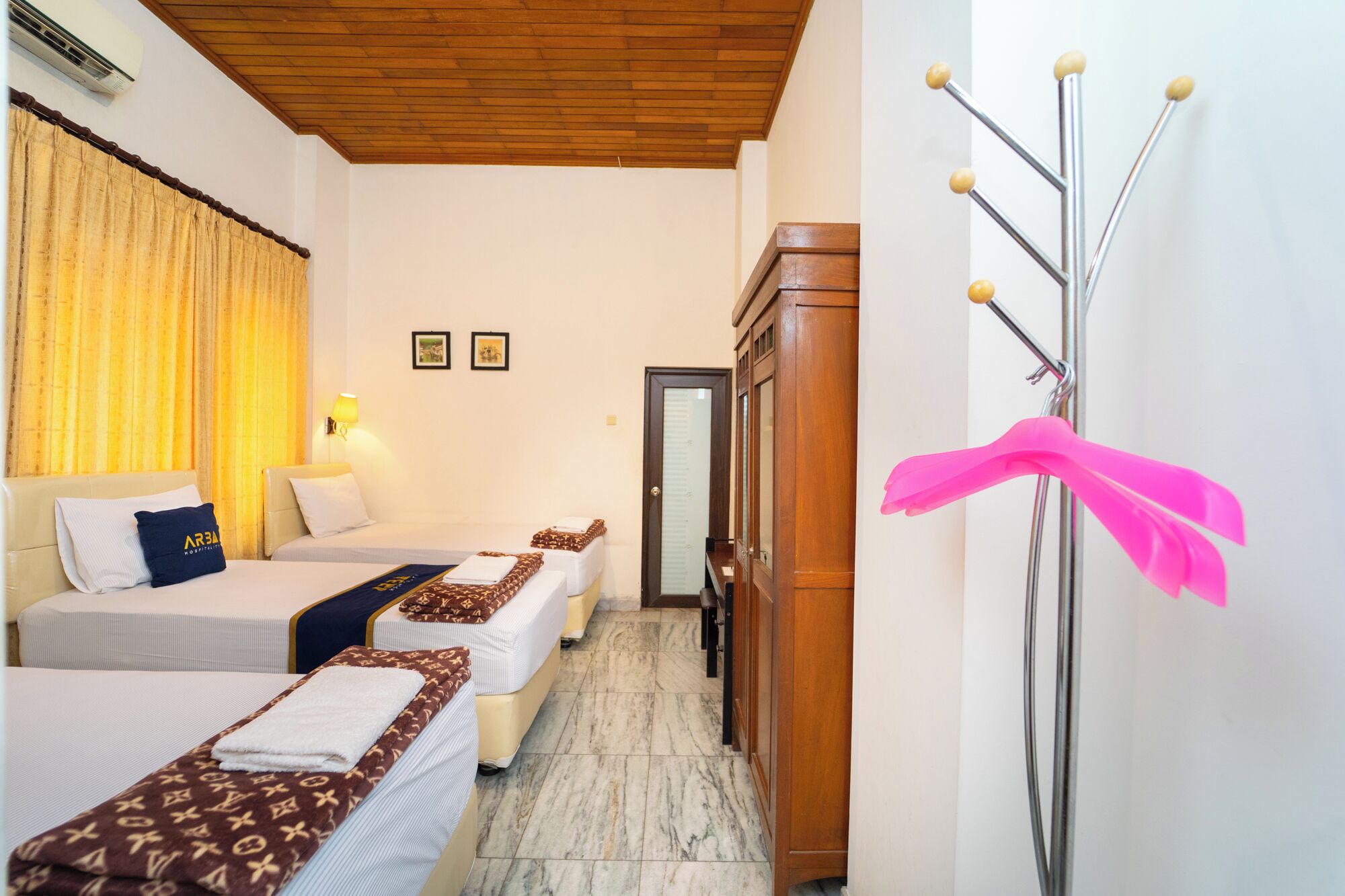 A hotel room with two beds, a wooden wardrobe, a mirror, and a pink decorative ribbon.
