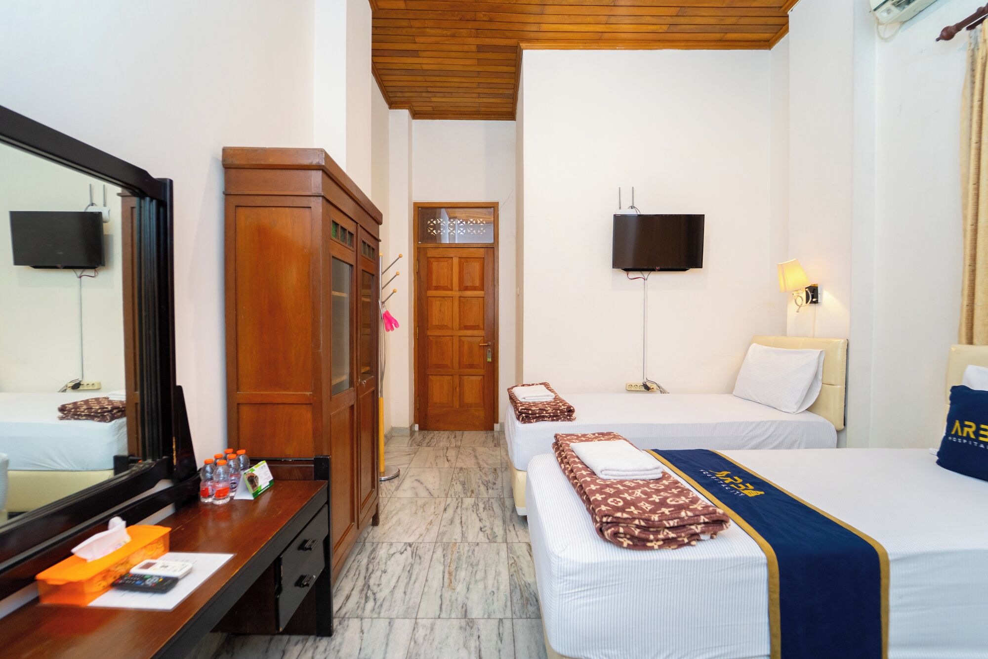 A hotel room with a wooden desk, two beds, a mirror, a TV, and a door.