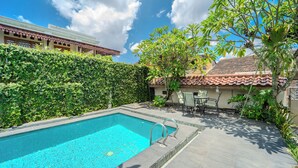 Una piscina con borde de baldosas, rodeada de vegetación y un edificio con techo de tejas.