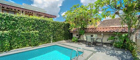 A swimming pool with a tiled deck, surrounded by greenery and a building with a tiled roof.