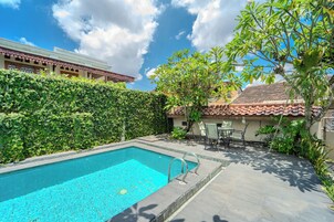 A swimming pool with a tiled deck, surrounded by greenery and a building with a tiled roof.