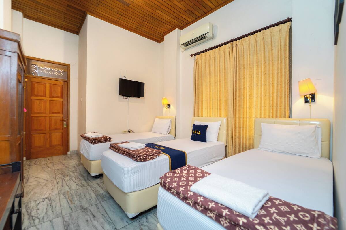 A hotel room with two beds, a wooden door, a television, an air conditioning unit, and a large window with curtains.