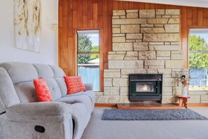 Ein Wohnzimmer mit Kamin aus Stein, einer grauen Couch mit roten Kissen und Blick ins Freie durch ein Fenster.