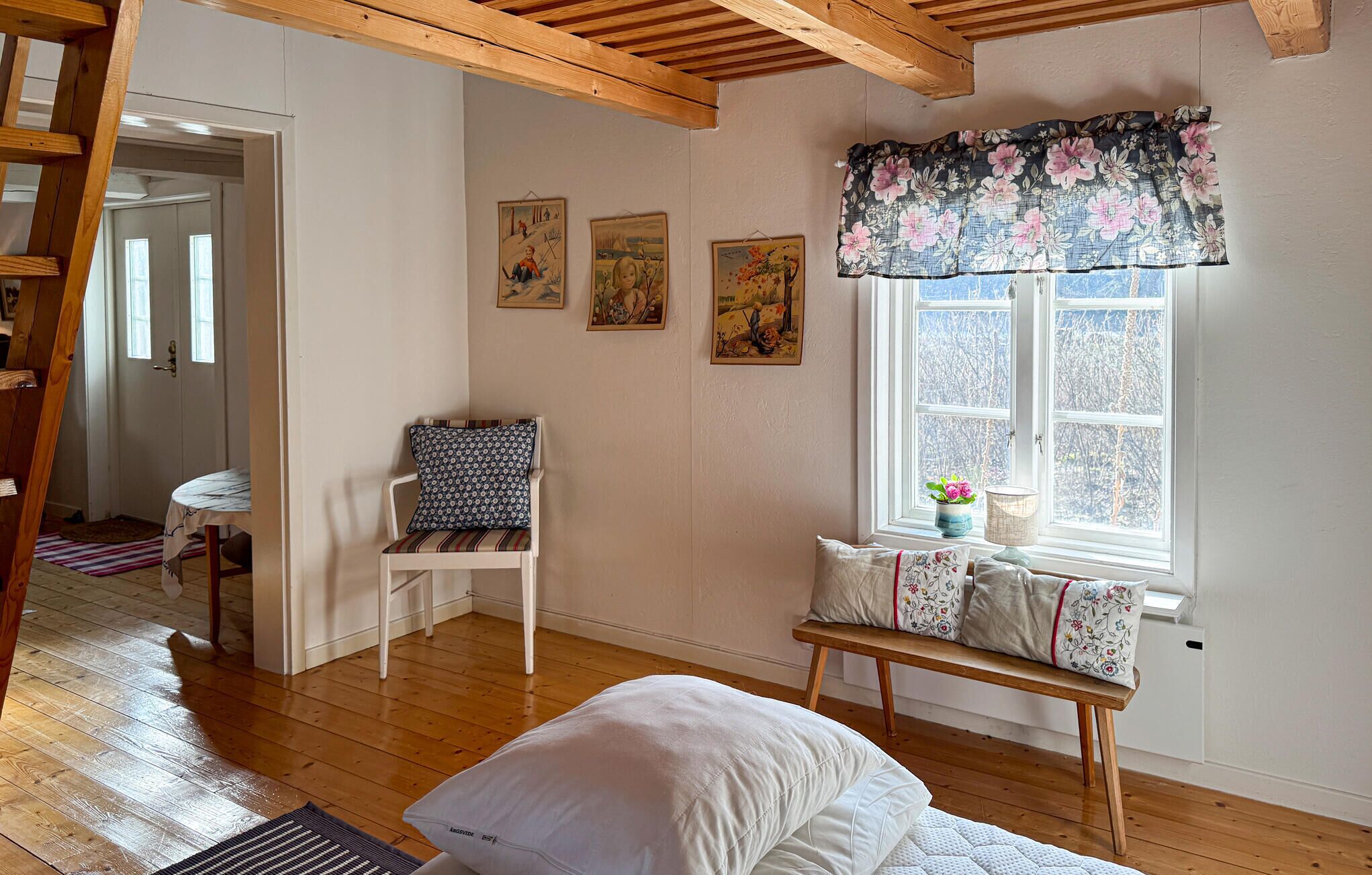 Una stanza con pavimento in legno, una sedia bianca con cuscino decorato, una panca con cuscini floreali e una finestra con tenda a fiori.