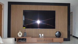 Ein modernes Wohnzimmer mit einem großen Flachbildfernseher, der an einer Wand aus Holzpaneelen befestigt ist, einer Holzunterhaltungsecke darunter und einem weißen Sofa auf der linken Seite.