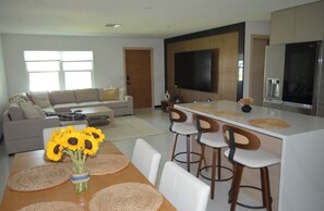 A modern living room with a sofa, a dining area with a table and chairs, a kitchen with a large island and bar stools, and a TV mounted on the wall. - Modern Coastal Getaway w/ Pool & Spacious Outdoor Living, 2 bed 2 bath (Port Charlotte)