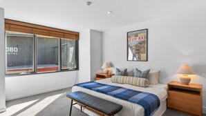 A bedroom with a large window, a bed with a blue checkered blanket, two bedside tables with lamps, and a poster on the wall. - Squatters Run 8: central ski-view stay w/ parking (Thredbo)