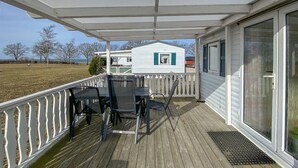 Een overdekt terras met een tafel en stoelen, een wit huis op de achtergrond en een grasveld.