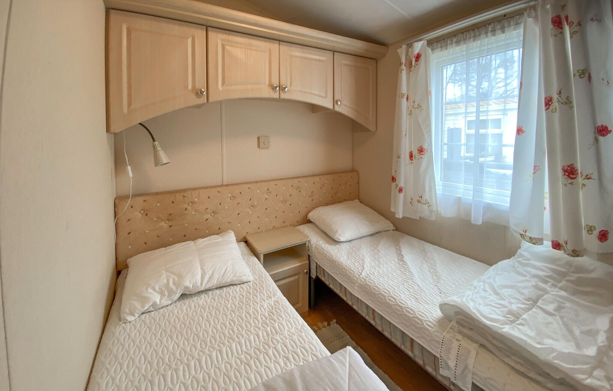 A small, single-room accommodation with two beds, a nightstand, a lamp, and a window with floral curtains.