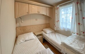 Een kleine, eenpersoonskamer met twee bedden, een nachtkastje, een lamp en een raam met bloemen gordijnen.