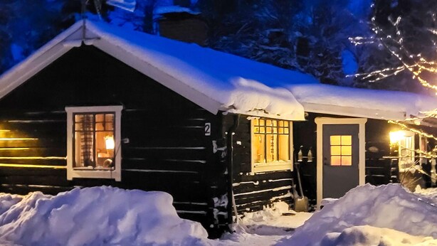 Une cabane dont le toit est recouvert de neige, avec une fenêtre illuminée et une porte.