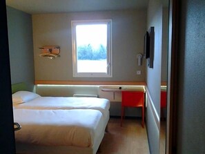 Ein Zimmer mit zwei Betten, einem Schreibtisch, einem Fenster und einem an der Wand befestigten Fernseher.