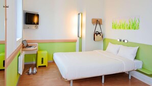 Una camera d'albergo con un letto, una scrivania con un portatile, una TV, una lampada a parete, un piccolo sgabello e un'opera d'arte appesa al muro.
