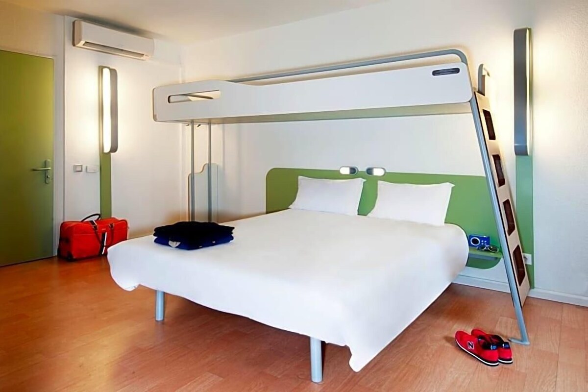 A bunk bed with a white mattress and green headboard, a red suitcase, and a pair of red slippers on the floor.