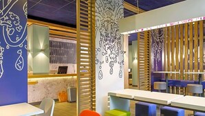 A modern interior with tables and colorful seating, a counter with a display, and decorative wall art. - Ibis Budget Givet (Givet)