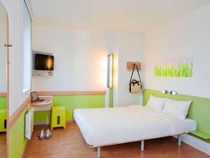 Une chambre d’hôtel comprenant un lit, un bureau avec un ordinateur portable, une télévision, une lampe fixée au mur, un petit tabouret et une œuvre d’art décorative.