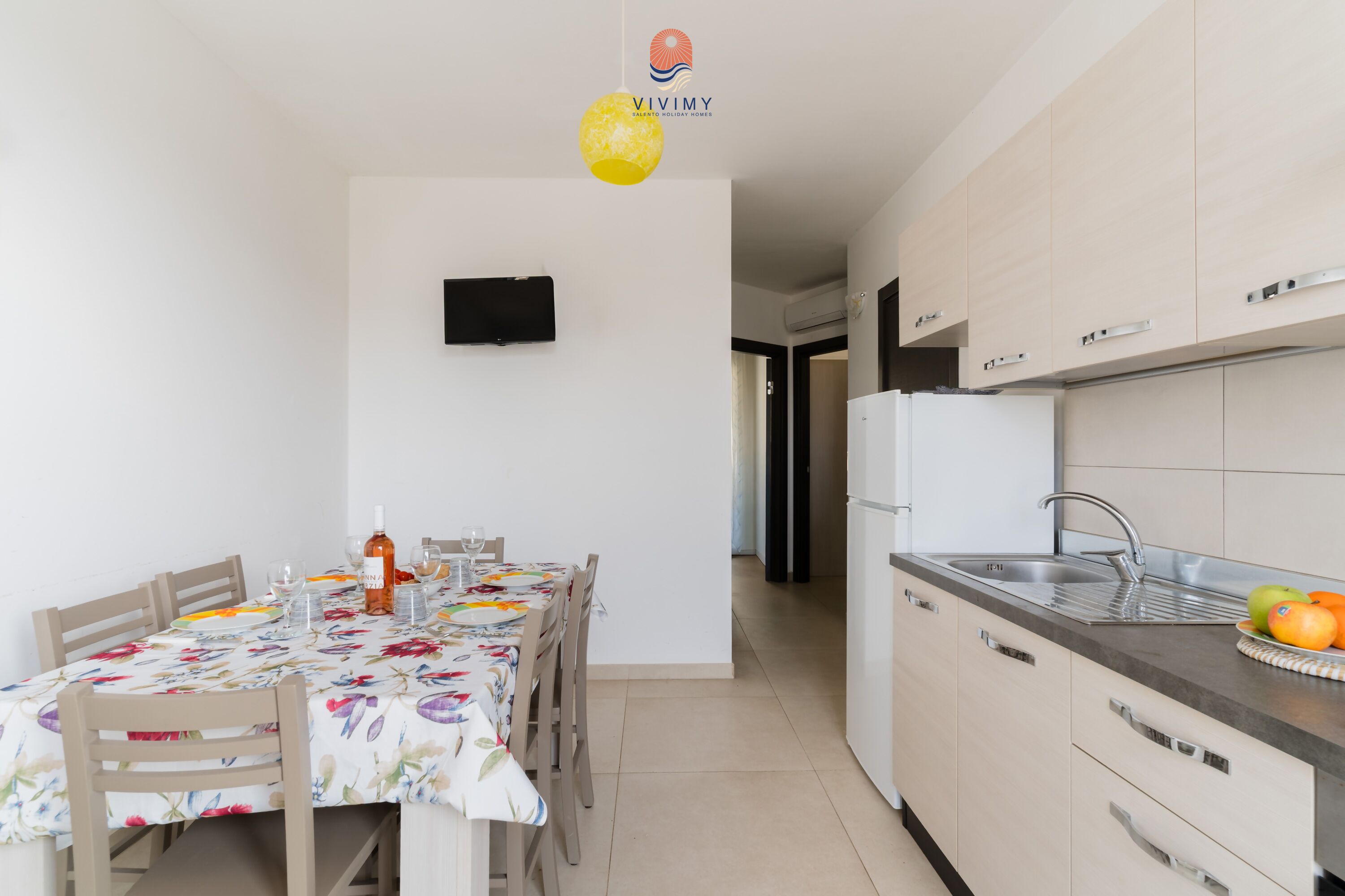 A modern kitchen with a dining area, a flat-screen TV, and a refrigerator.
