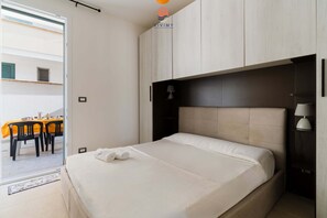 Ein Schlafzimmer mit einem Bett, einem Nachttisch, einer Lampe und Blick auf einen Außenbereich mit Essplatz.