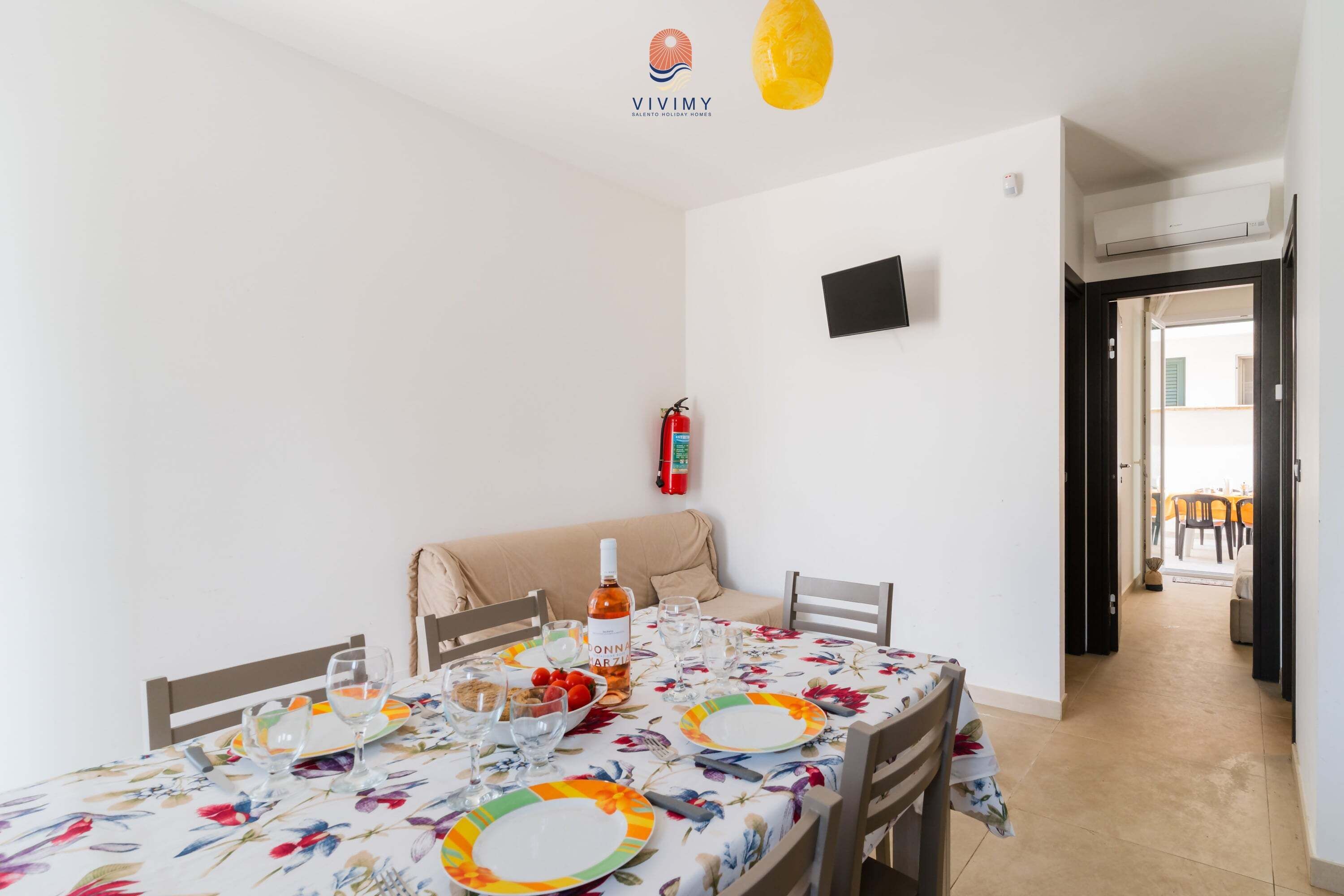 A dining area with a table set for four, a sofa, a fire extinguisher, and an open door leading to another room.