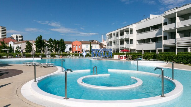 Una piscina de diseño circular, rodeada de sillones de playa y sombrillas, con un edificio de varios pisos al fondo.