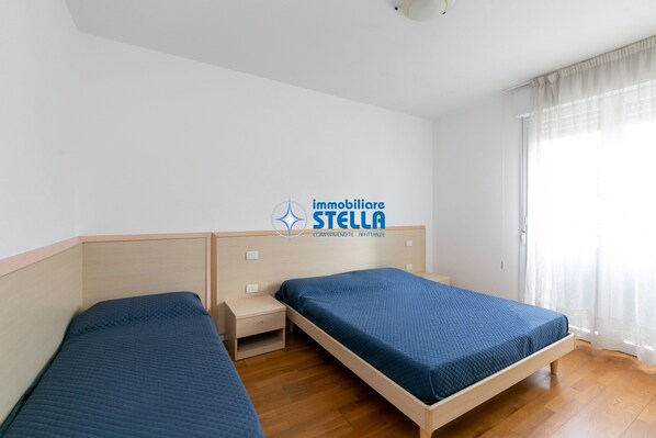 Een hotelkamer met twee enkele bedden, houten hoofdeinden, een klein nachtkastje en een raam met dunne gordijnen.