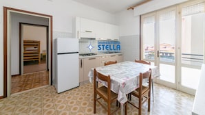 A kitchen with a dining table, a refrigerator, and a view through the sliding glass door. - Romeo [bilocale+parking] (Lido di Jesolo)