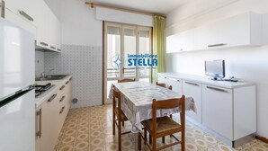 A compact kitchen with a dining area, a TV, and a window with curtains. - Romeo [bilocale+parking] (Lido di Jesolo)