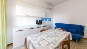 A room with a dining table, chairs, a blue sofa, a white cabinet, and an air conditioning unit. - Romeo [bilocale+parking] (Lido di Jesolo)