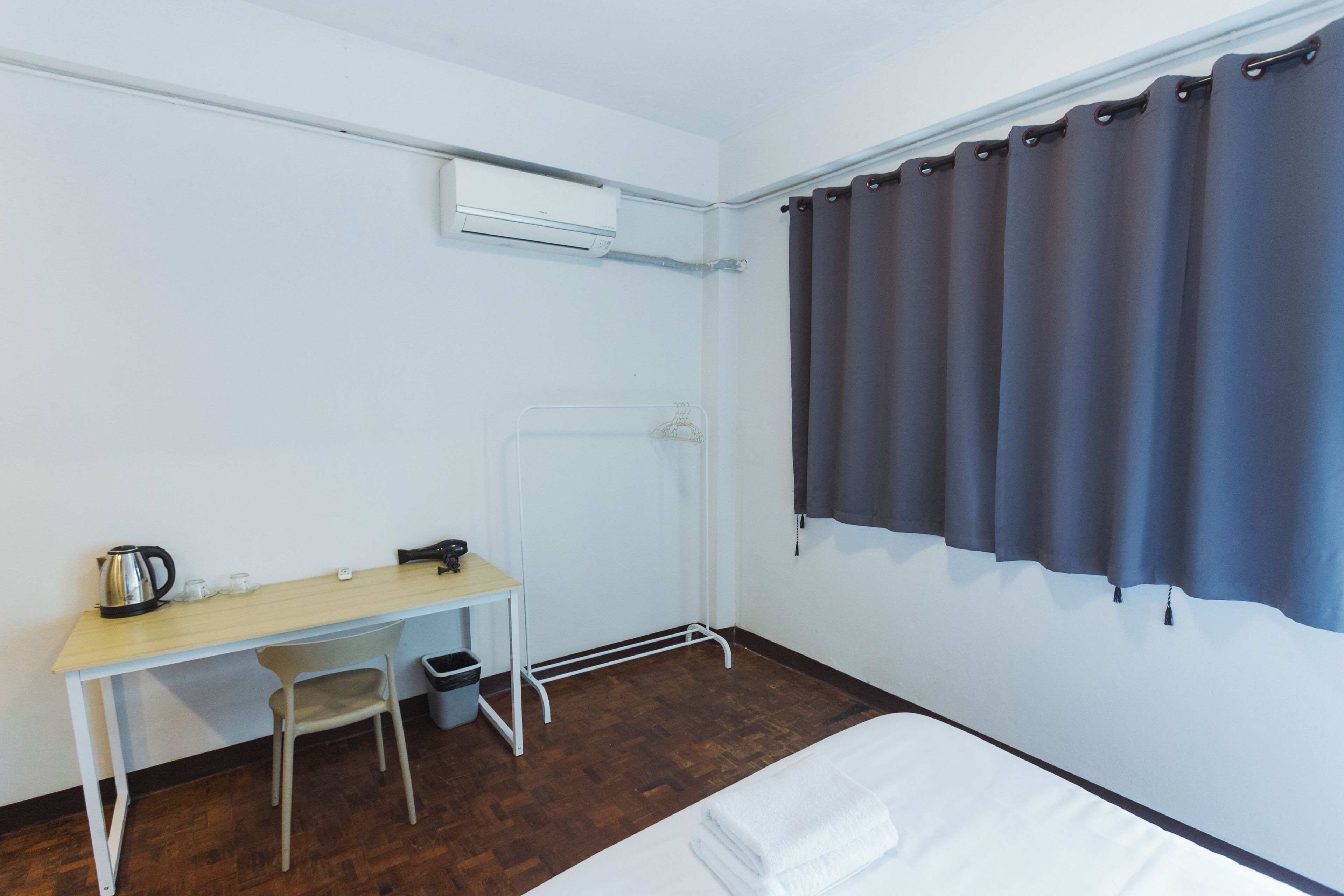 Una habitación con una cama, un escritorio, una silla, una mesita con una tetera y un cubo de basura.