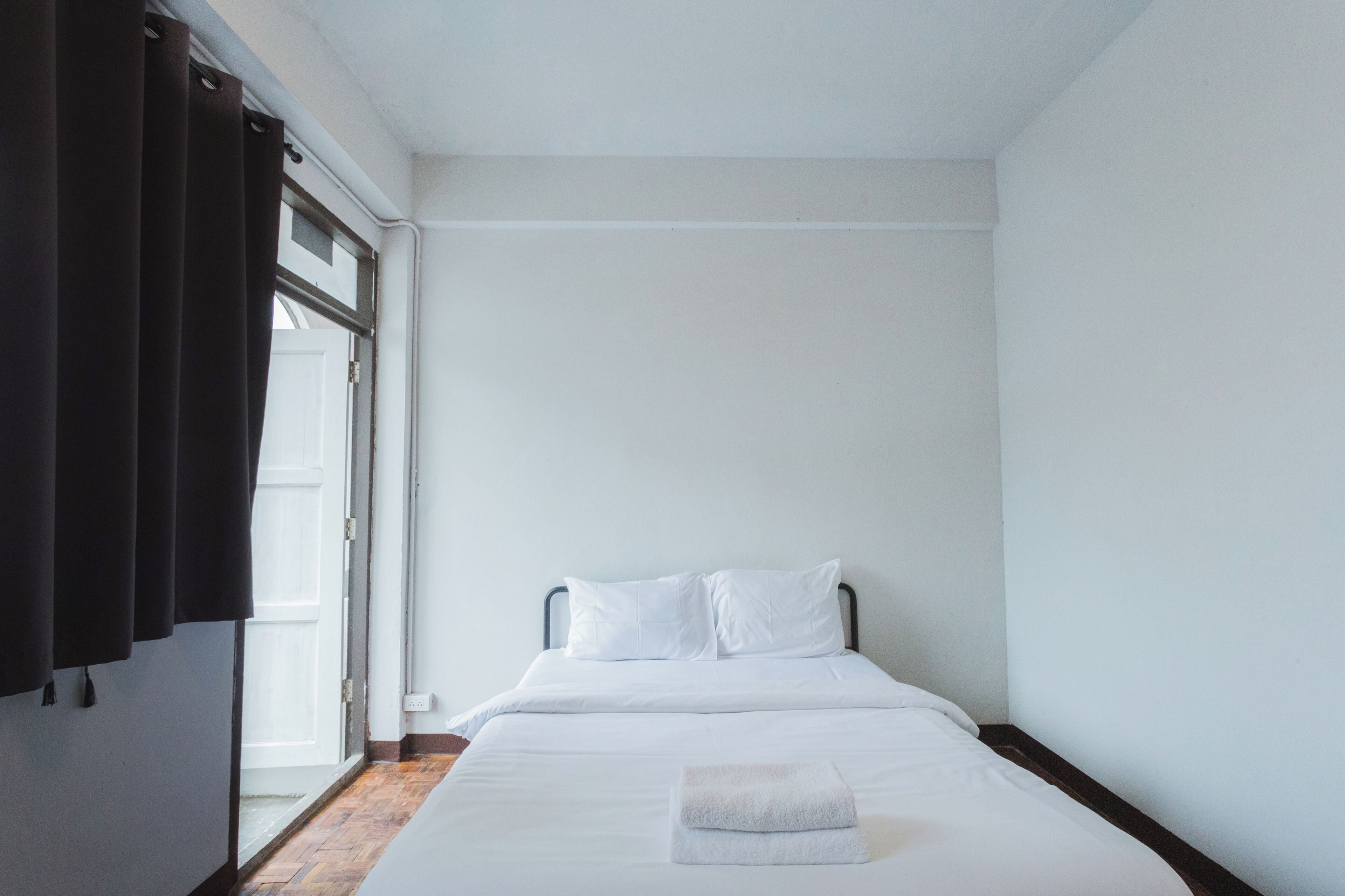 A bedroom with a bed, white bedding, a folded towel, a window with curtains, and a wooden floor.