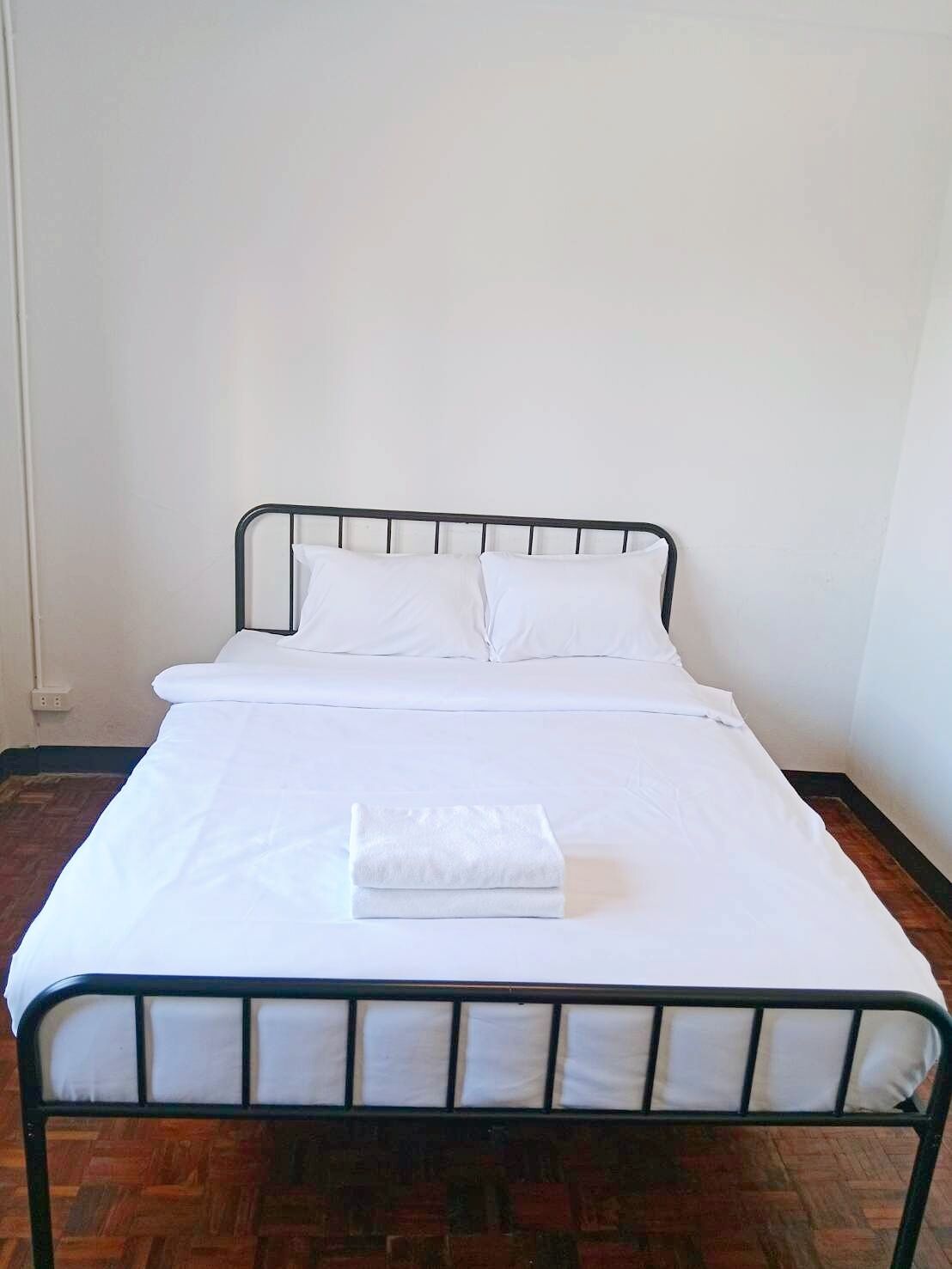 Una cama individual con estructura metálica, ropa de cama blanca y una toalla blanca doblada sobre la cama.