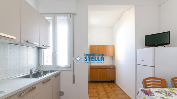Eine kleine Küche mit Fenster, Spüle, Schränken, einem Holzschrank, einem Tisch mit Stühlen und einem Fernseher auf einem Standfuß.