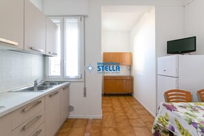 Une cuisine de taille réduite, avec une fenêtre, un évier, des placards, un meuble en bois, une table avec des chaises et un téléviseur posé sur un meuble.
