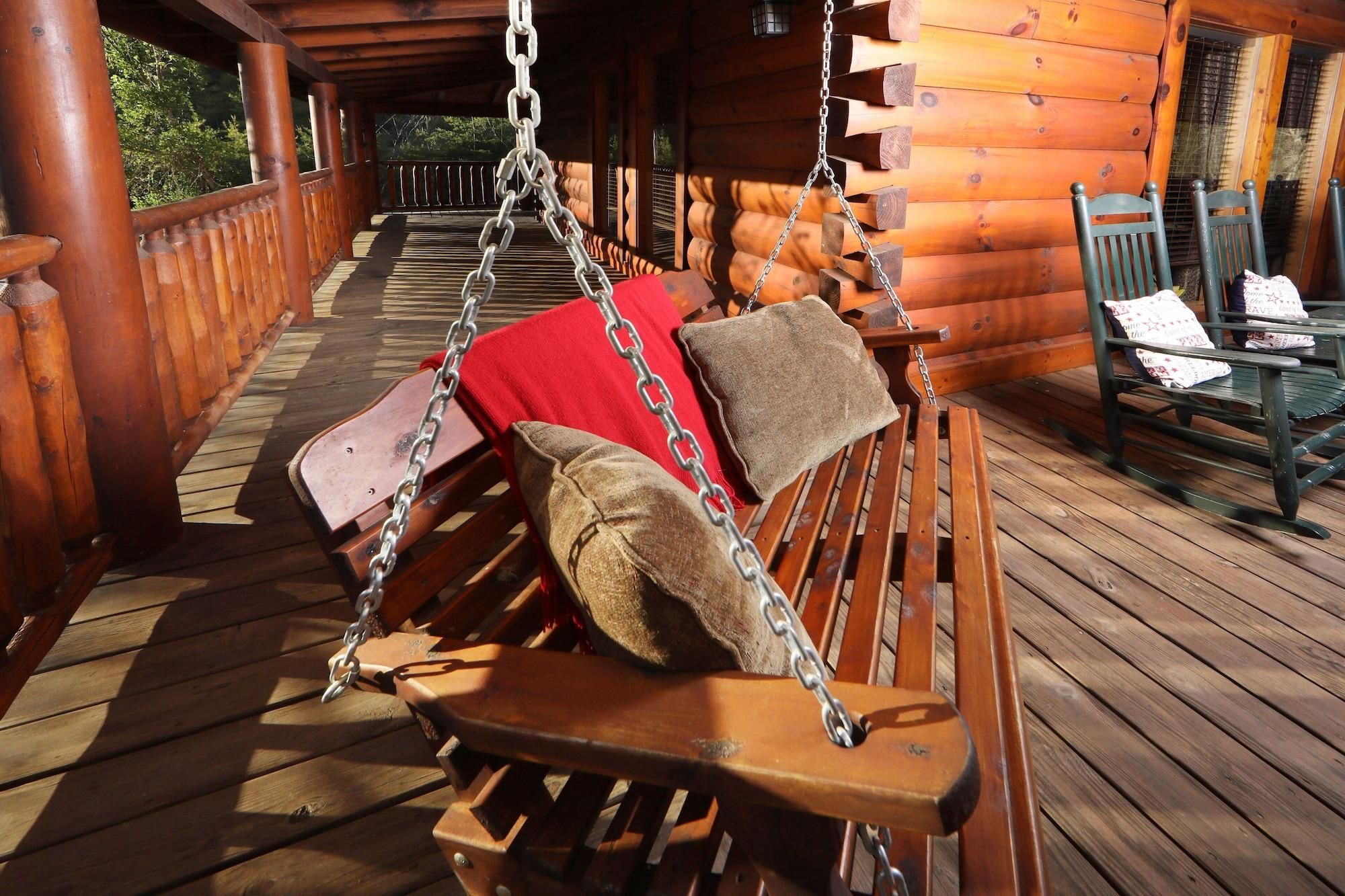 Une chaise suspendue en bois avec des coussins et une chaîne, un fauteuil à bascule et une terrasse en bois offrant une vue sur les arbres.