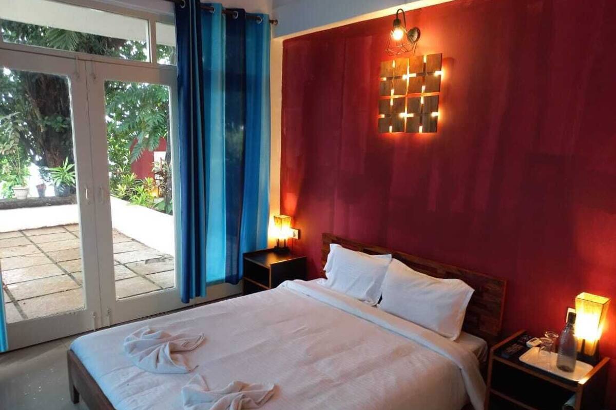 A hotel room with a bed, two bedside tables, a nightstand, a window with curtains, and a view of the outdoors.