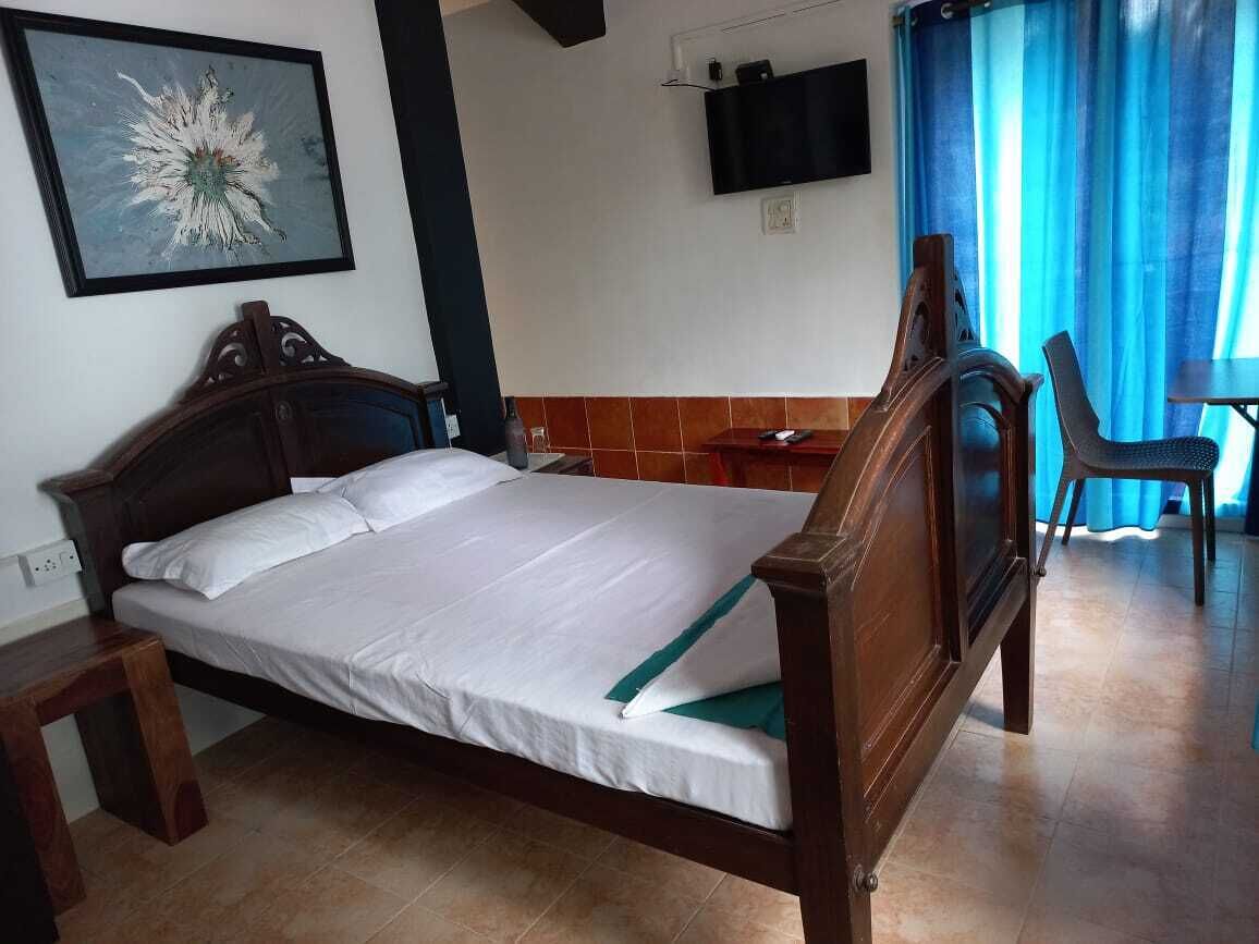 Una cama bien hecha con cabecera de madera, una mesita de noche pequeña, un televisor montado en la pared y una cortina azul.