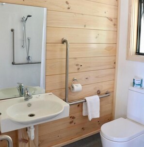 A bathroom with a wooden wall, a white sink, a mirror, a shower, and a toilet. - Lakes Edge Lodge (Lake Tekapo)