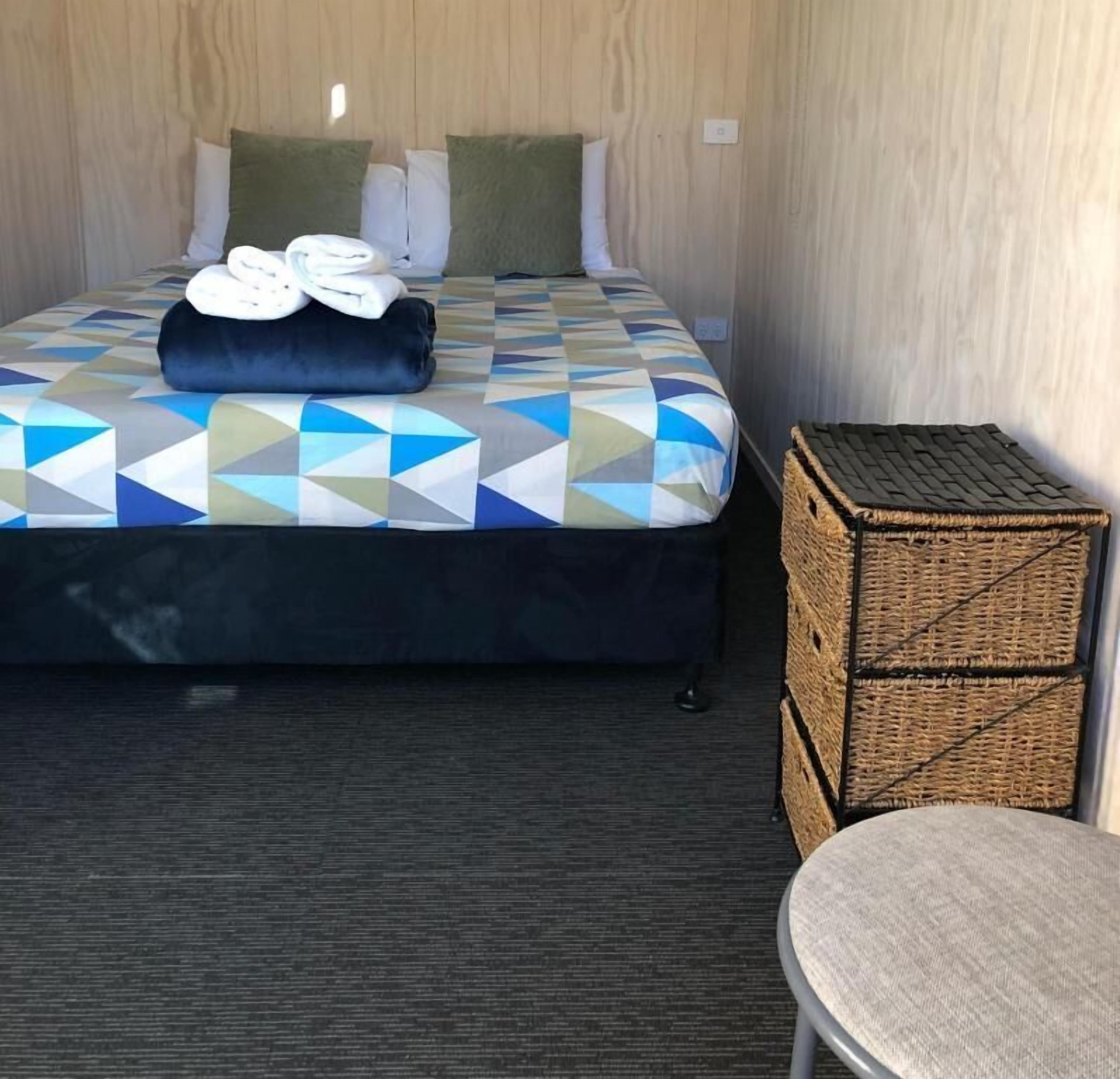 A hotel room with a bed, pillows, a folded towel, a small wicker basket, and a chair.