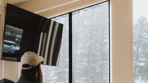 A person sitting on a bench inside a room with a television mounted on the wall, a window with a view of trees and a parked car outside. - Lakes Edge Lodge (Lake Tekapo)