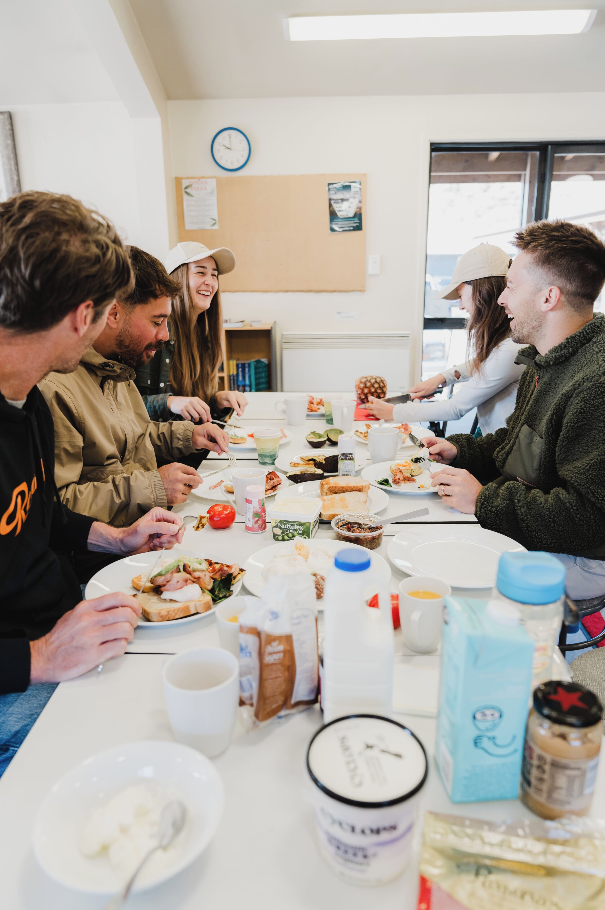 Un grupo de personas sentadas alrededor de una mesa con comida y bebidas, en una habitación con un reloj en la pared.