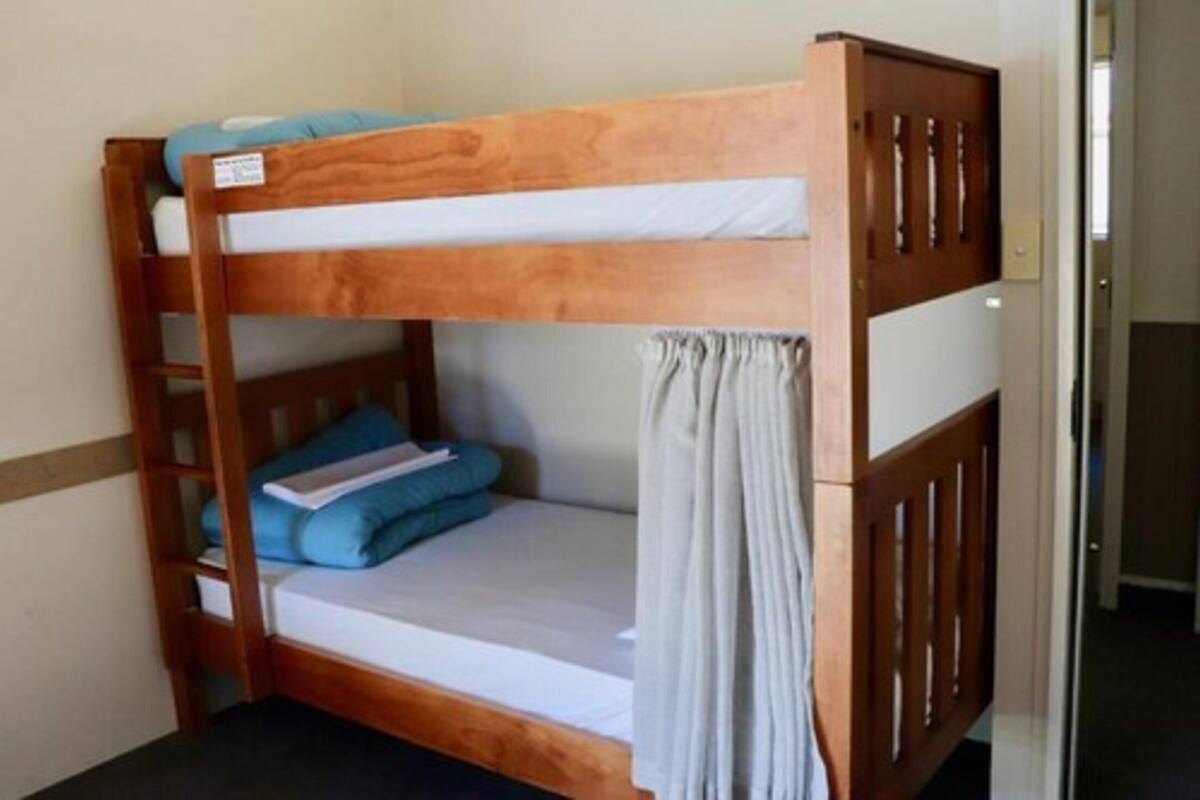 A bunk bed with a wooden frame, a lower bunk with a grey mattress and a white sheet, and a top bunk with a blue mattress and pillows.