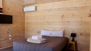 A room with wooden walls, a bed with a quilted mattress, a flat-screen TV mounted on the wall, and a small table with a lamp. - Lakes Edge Lodge (Lake Tekapo)
