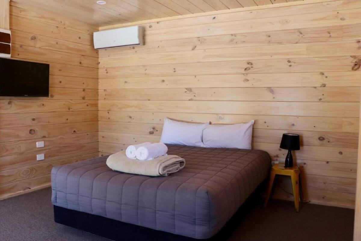 A room with wooden walls, a bed with a quilted mattress, a flat-screen TV mounted on the wall, and a small table with a lamp.