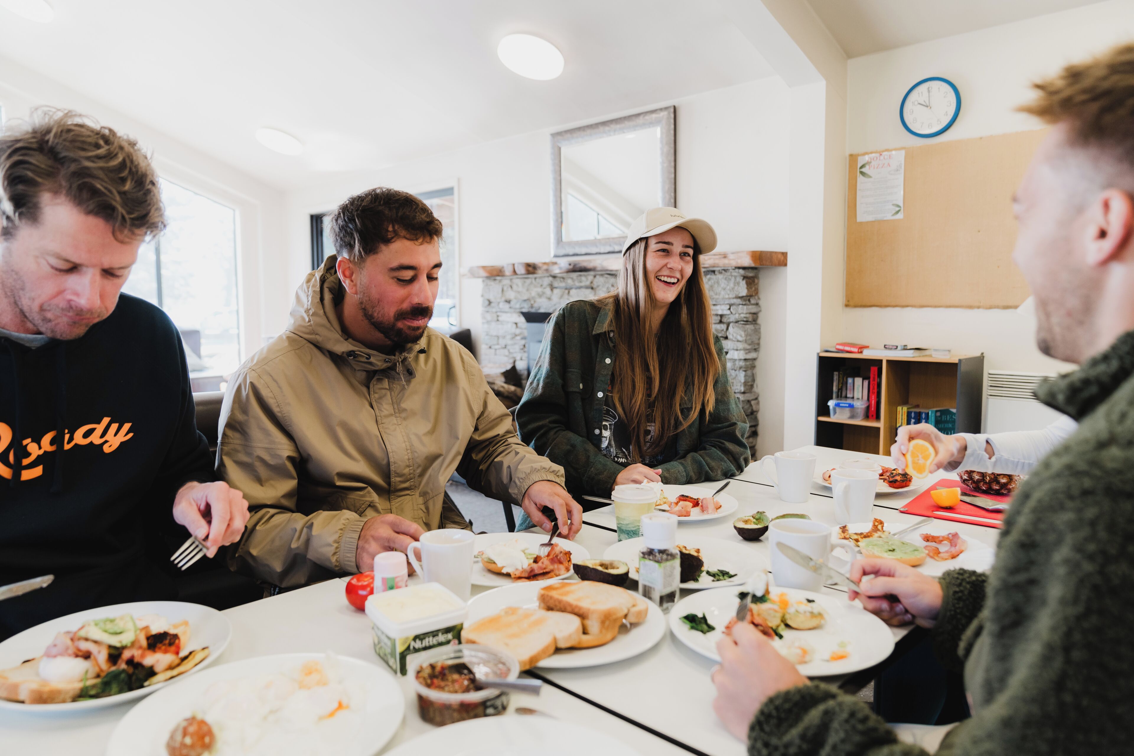 Un grupo de personas cenando en una mesa con varios platos, un reloj en la pared y una estantería al fondo.
