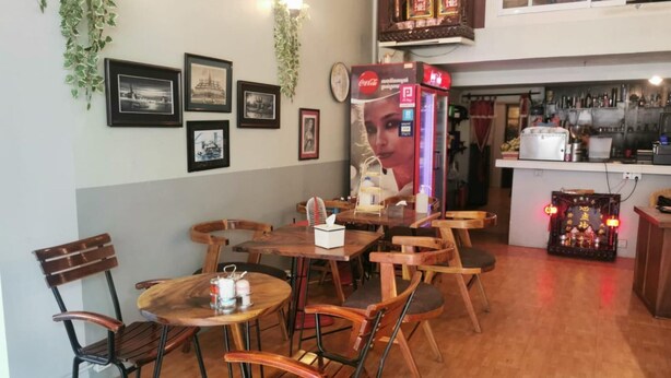 A restaurant interior with wooden tables and chairs, a Coca-Cola vending machine, framed pictures on the wall, and a kitchen area in the background. - Sovanphum Riverside Hotel (Phnom Penh)