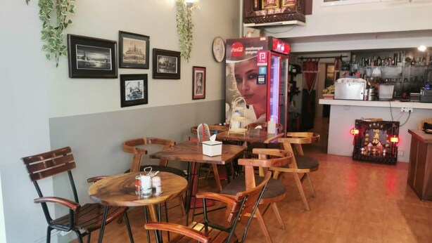 A restaurant interior with wooden tables and chairs, a Coca-Cola vending machine, framed pictures on the wall, and a kitchen area in the background. - Sovanphum Riverside Hotel (Phnom Penh)