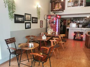 A restaurant interior with wooden tables and chairs, a Coca-Cola vending machine, framed pictures on the wall, and a kitchen area in the background. - Sovanphum Riverside Hotel (Phnom Penh)